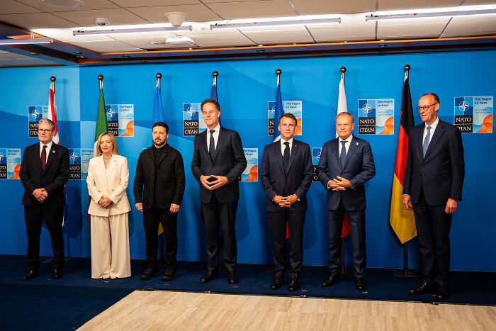 Group of political leaders standing in front of NATO and national flags discussing Ukraine war and Zelenskyy&rsquo;s role.