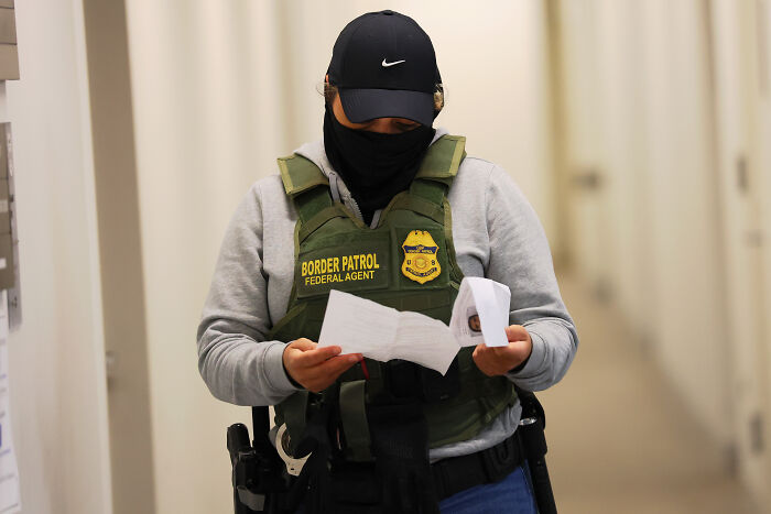 Immigration officer wearing tactical gear and reading documents while standing in a hallway near LA school detention area.
