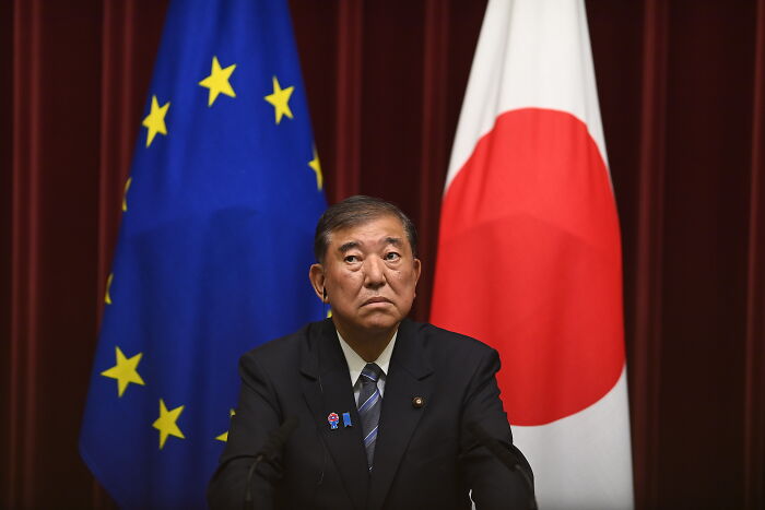 Japanese official in a dark suit speaking at a podium with European Union and Japan flags behind on avoiding World War III risks.