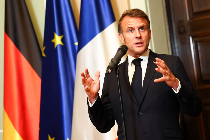 Man speaking at a podium with French, German, and EU flags behind, related to French cops clashing with dad and son on UK-bound boat.