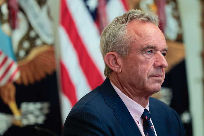 Robert F. Kennedy Jr. wearing a suit, seated with American flags in the background amid MAHA uproar discussion.