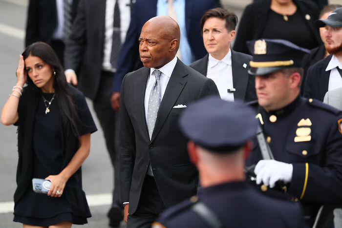 Eric Adams walking in a suit, surrounded by police officers and people, amid NYC mayoral race tensions about adult work decriminalization.