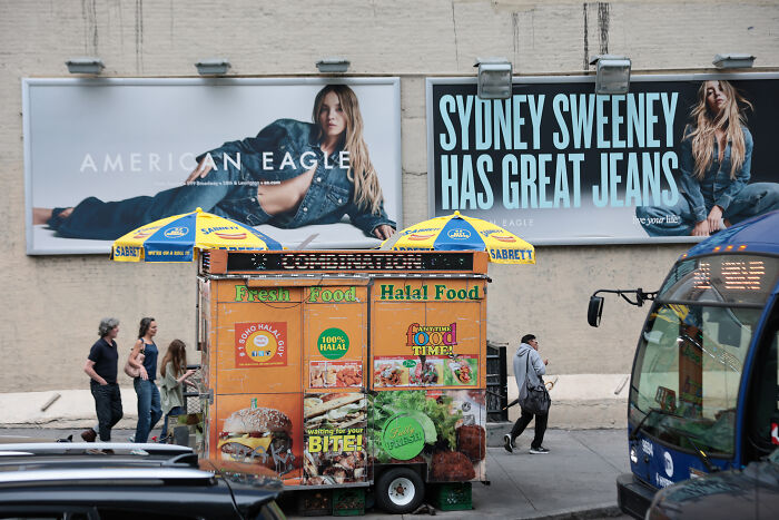 Sydney Sweeney in controversial jeans advert displayed on large billboards in urban street setting with food cart and pedestrians.