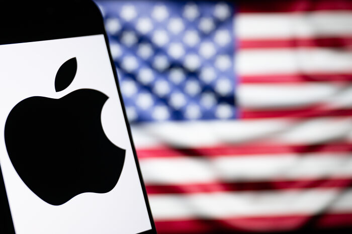 Apple logo displayed on a smartphone screen with a blurred American flag in the background representing Apple&rsquo;s Tim Cook and White House.