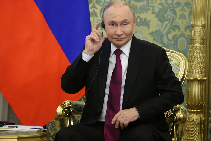 Vladimir Putin sitting on ornate chair wearing black suit and red tie with Russian flag in the background before high-stakes meeting.