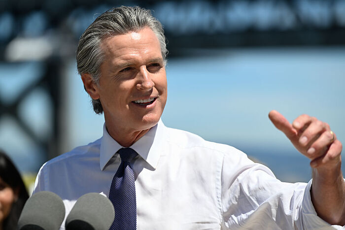 Gavin Newsom speaking outdoors near microphones, smiling and gesturing with his hand in front of a blurred background.
