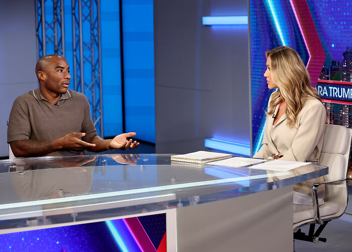 Charlamagne Tha God speaking during an interview in a studio setting with a female host at a glass table.