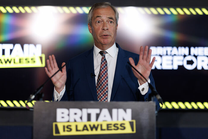 Man in suit speaking at podium with Britain is lawless sign, highlighting French cops clashing with dad and son boarding UK-bound boat.