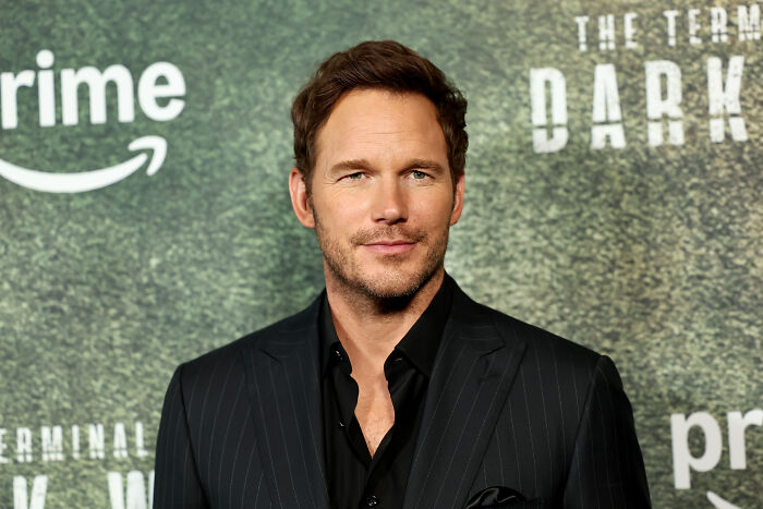 Chris Pratt posing at a premiere event, dressed in a black pinstripe suit with a green textured background.