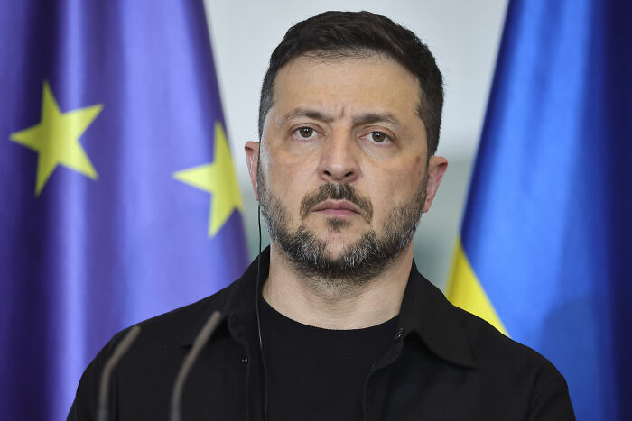 Ukrainian leader standing solemnly with European Union and Ukraine flags, symbolizing West Bank-style occupation of Ukraine.