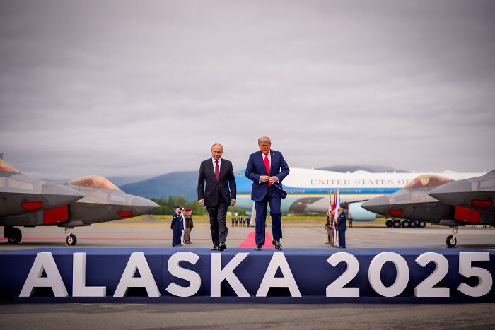 Donald Trump and Vladimir Putin walking at an airport with Alaska 2025 sign, related to Ukraine war discussion.