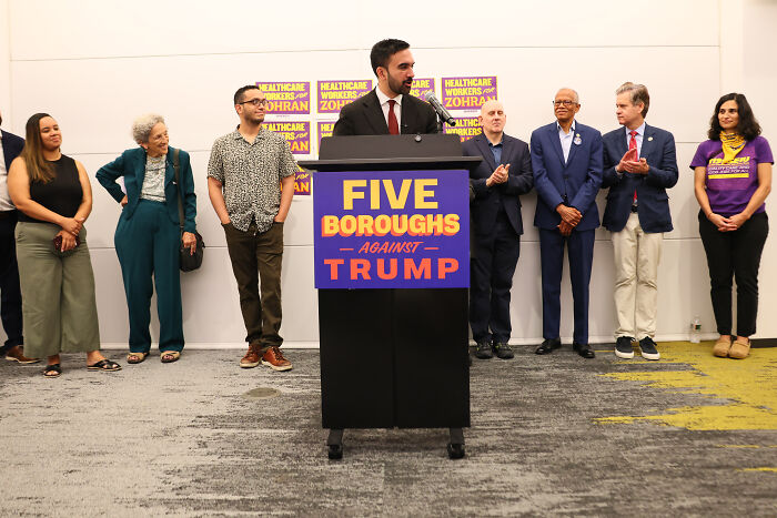 Zohran Mamdani speaking at a podium with supporters, addressing adult work decriminalization in NYC mayoral context.