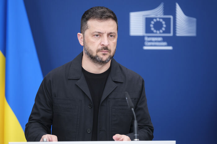 Volodymyr Zelenskyy speaking at a podium with Ukraine and EU flags, related to Ukraine war and Trump comments.