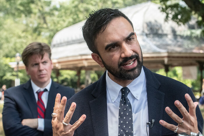 Zohran Mamdani speaking passionately outdoors, with a blurred man in a suit behind him, NYC mayoral hopeful discussion.