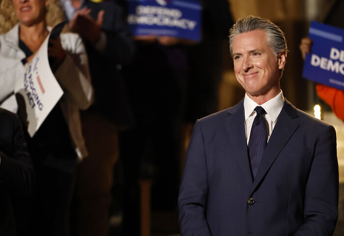 Gavin Newsom smiling in a suit at an event with supporters holding democracy signs, related to trolling JD Vance viral photo.