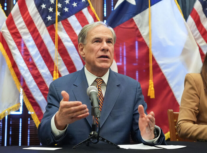 Texas GOP leader in a suit speaking at a press conference with American and Texas flags behind him on partisan map.