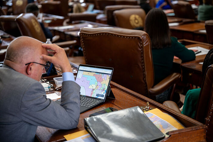Man examining a political map on a laptop in a legislative chamber during Texas GOP redistricting discussions.
