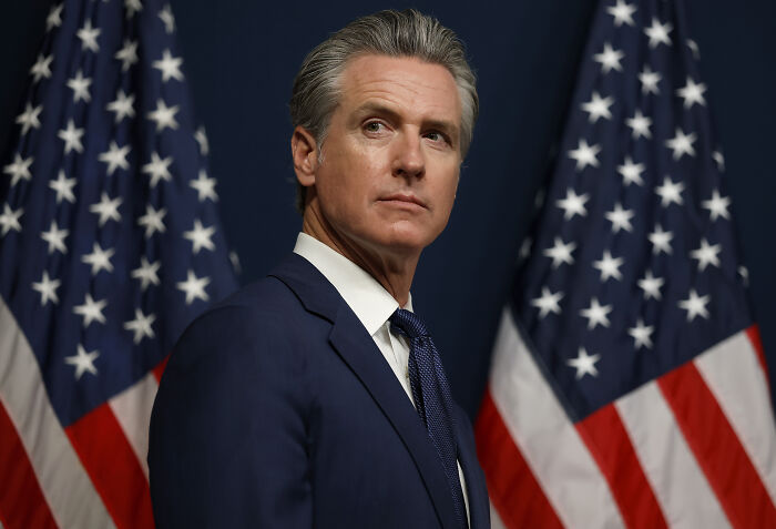 Gavin Newsom in a suit standing between American flags during a formal event, showcasing his troll game edge.