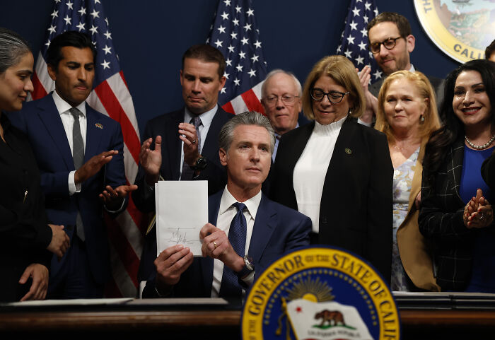 California Governor signing the redistricting plan, surrounded by officials and U.S. flags in a formal setting.