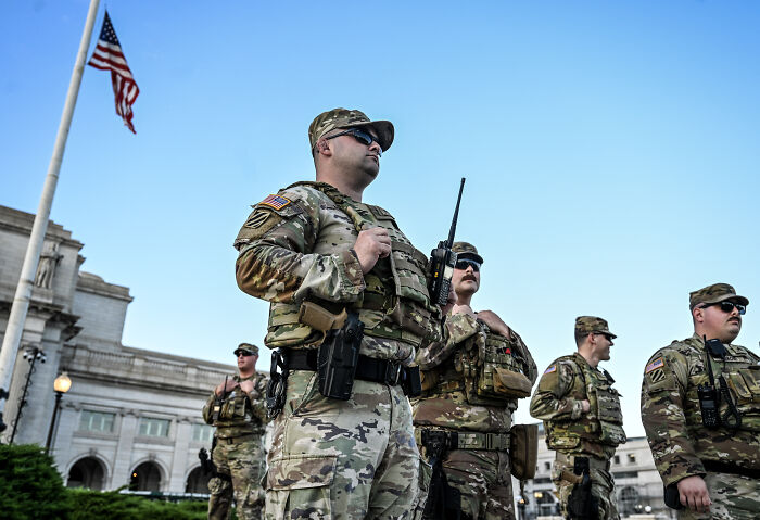 National Guard members in Washington D.C. wearing camouflage uniforms standing near a flagpole during daylight.