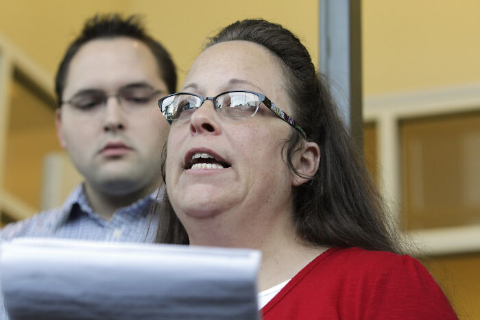 Woman wearing glasses and red shirt speaking, with man in background, illustrating Supreme Court same-sex marriage debate.
