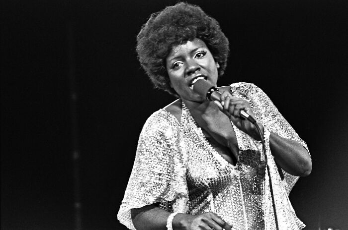 LGBTQ+ icon Gloria Gaynor singing on stage, wearing a sparkly top and holding a microphone passionately.