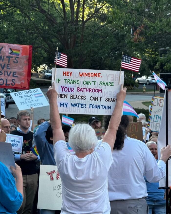 Protester at trans rights rally holding racist sign about water fountain, surrounded by crowd with flags and protest signs.
