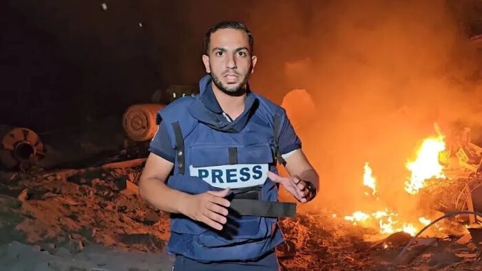 Journalist wearing a press vest reporting at night near burning debris, highlighting dangers to press in conflict zones.