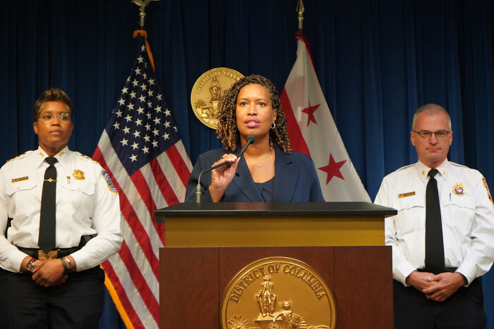 Woman speaking at podium with District of Columbia seal, flanked by police officers, representing federal takeover in Washington, D.C.