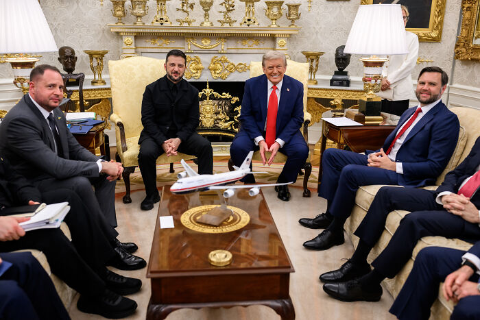 Volodymyr Zelenskyy in a black suit at a White House meeting with Donald Trump and officials seated around a table.