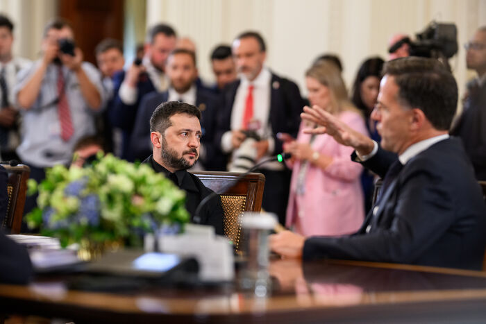 Volodymyr Zelenskyy in black suit attends White House meeting with officials and media in the background.