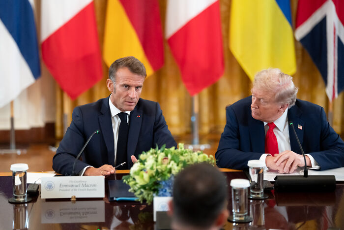 Two men in suits at a formal meeting table with flags in the background, focusing on Zelenskyy and Trump discussion.