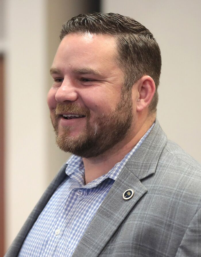 GOP lawmaker with beard and plaid shirt smiling indoors amid child exploitation video charge and resignation news.