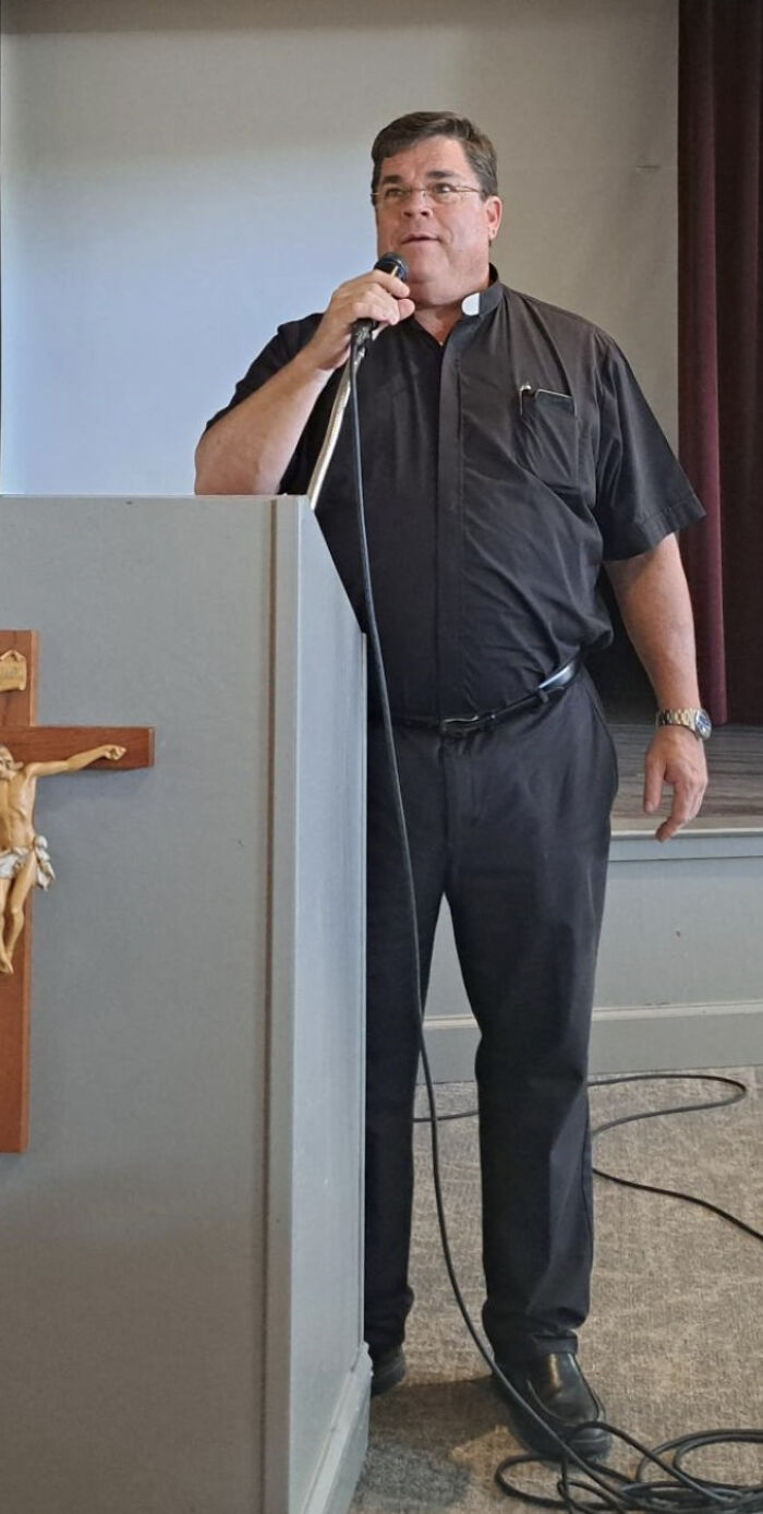 Alabama priest in black clerical shirt speaking at podium with crucifix, microphone in hand during indoor event.