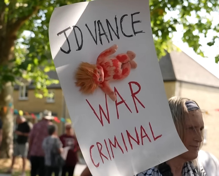 Protester holds sign calling JD Vance a war criminal amid villagers&rsquo; fury over disabled parking takeover.