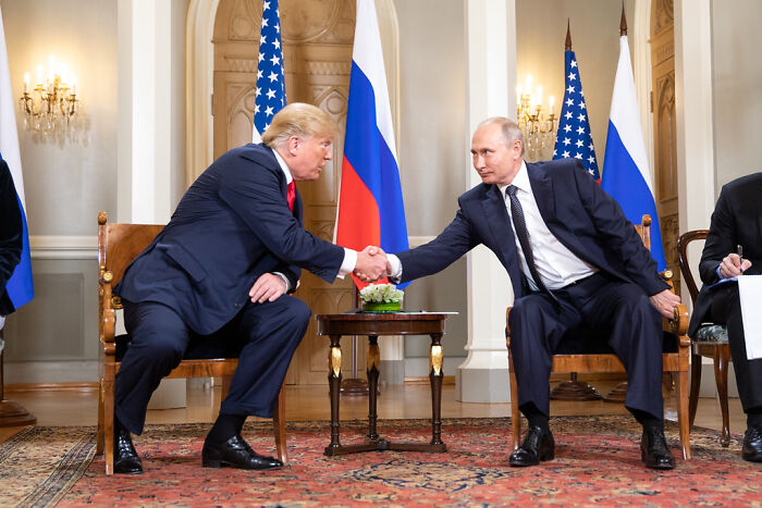 Donald Trump and Vladimir Putin shaking hands during a formal meeting with flags of the USA and Russia in the background.