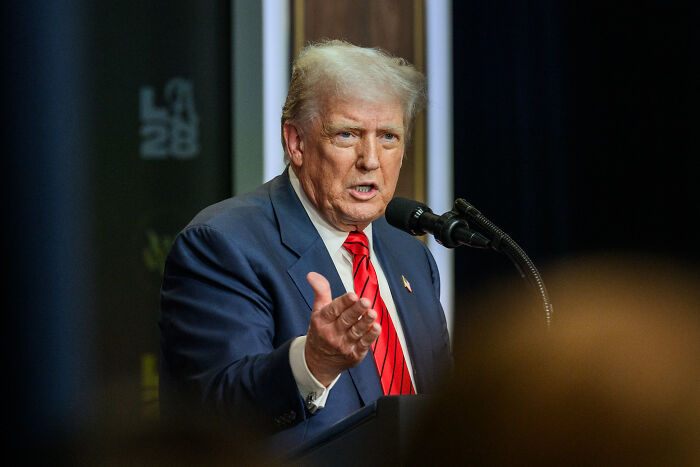 Donald Trump speaking at a podium wearing a blue suit and red tie as Jimmy Kimmel mocks Trump on-air.