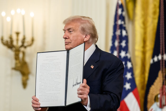 Donald Trump holding a signed document with American flags and a chandelier in the background in a formal setting