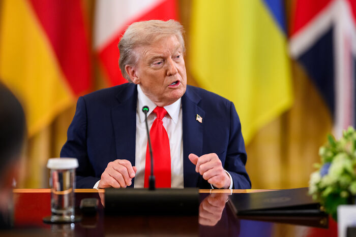 Former President Trump speaking at a conference table with international flags behind, amid tensions with Venezuela concerns.