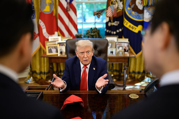 Donald Trump speaking in the Oval Office, with a red hat on the desk, amid controversy over domestic violence remarks.