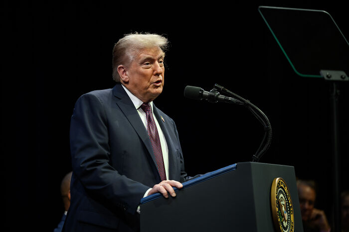 Donald Trump at a podium with presidential seal speaking into microphone during a public event on a dark stage.