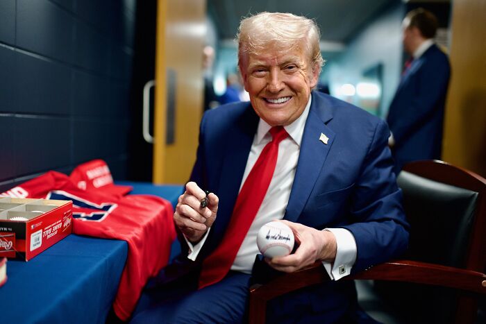 Donald Trump in a blue suit and red tie smiling while signing a baseball, linked to AI post and medbed conspiracy theory controversy.