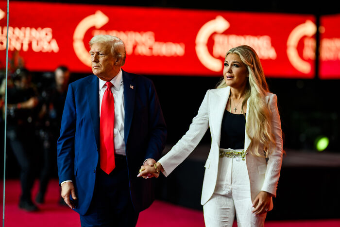 Donald Trump and a woman holding hands, attending an event with bright red background and logos.