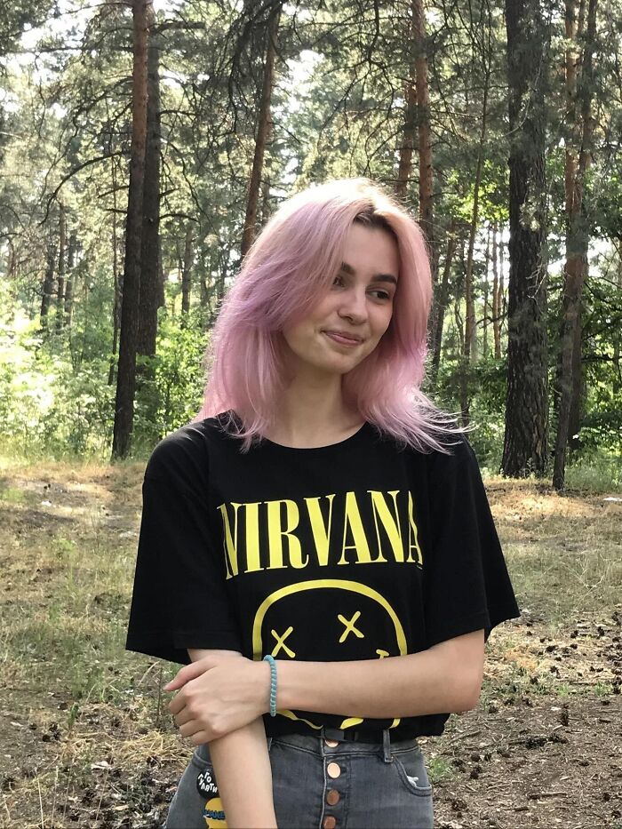 Young woman with pink hair wearing a Nirvana shirt standing in a forest, related to Ukrainian refugee stabbing incident.