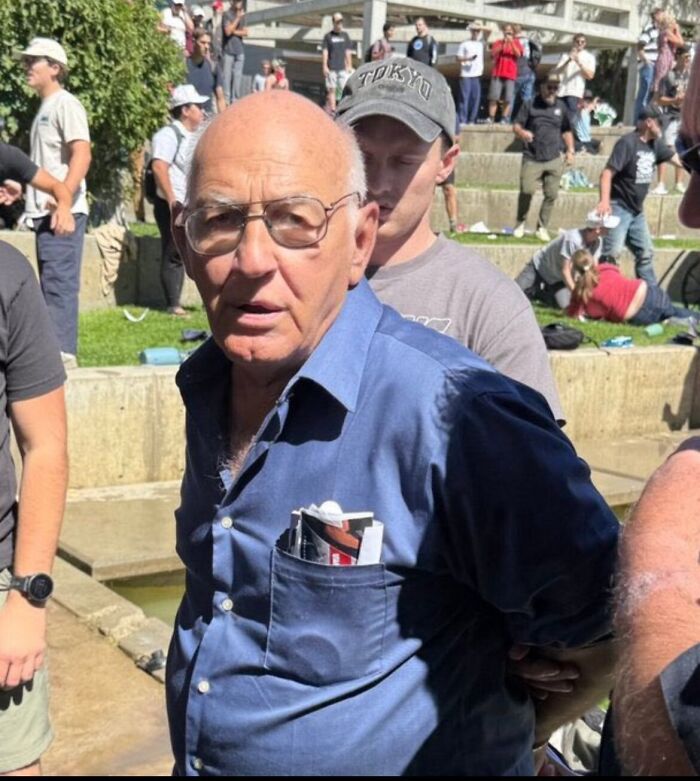 Elderly man in glasses and blue shirt surrounded by people discussing Charlie Kirk shooting conspiracy theories outdoors.