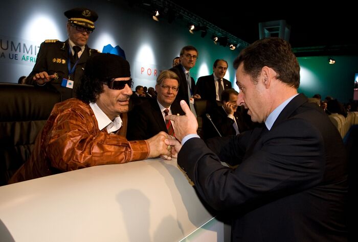 Former French President Nicolas Sarkozy in a dark suit talking with a man wearing sunglasses and a brown jacket at a summit event.