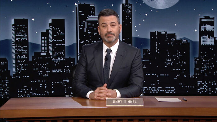 Jimmy Kimmel in a suit seated at a desk with a city skyline backdrop, related to Trump welcoming Kimmel pulled off air.