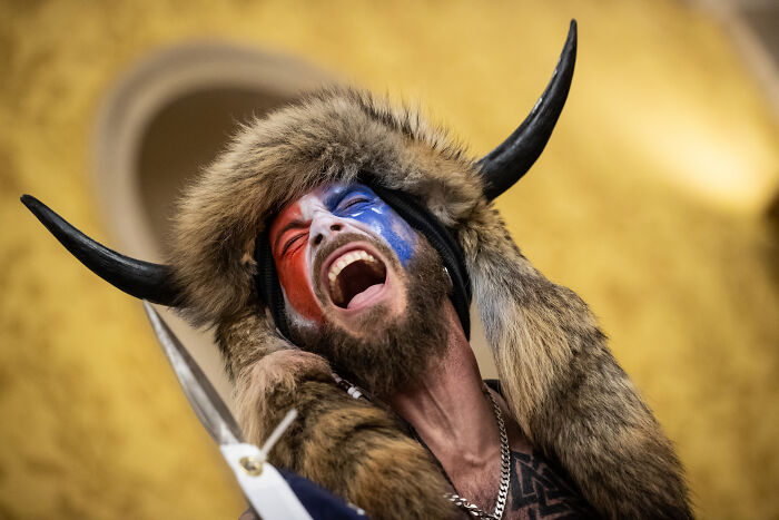 Man wearing horned fur hat with red and blue face paint shouting, representing QAnon Shaman in a political protest.