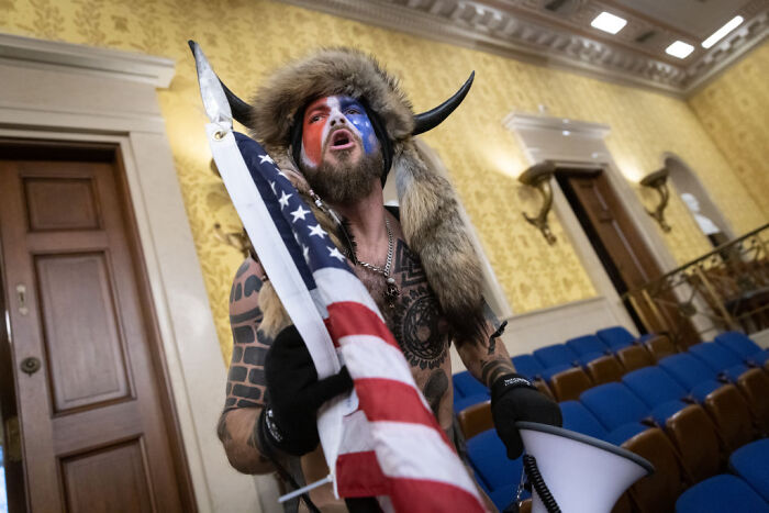 QAnon Shaman with face paint and fur hat holding American flag inside a government building.