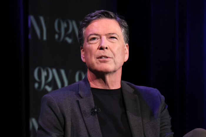 James Comey speaking at a public event, wearing a dark blazer and black shirt with a serious expression.